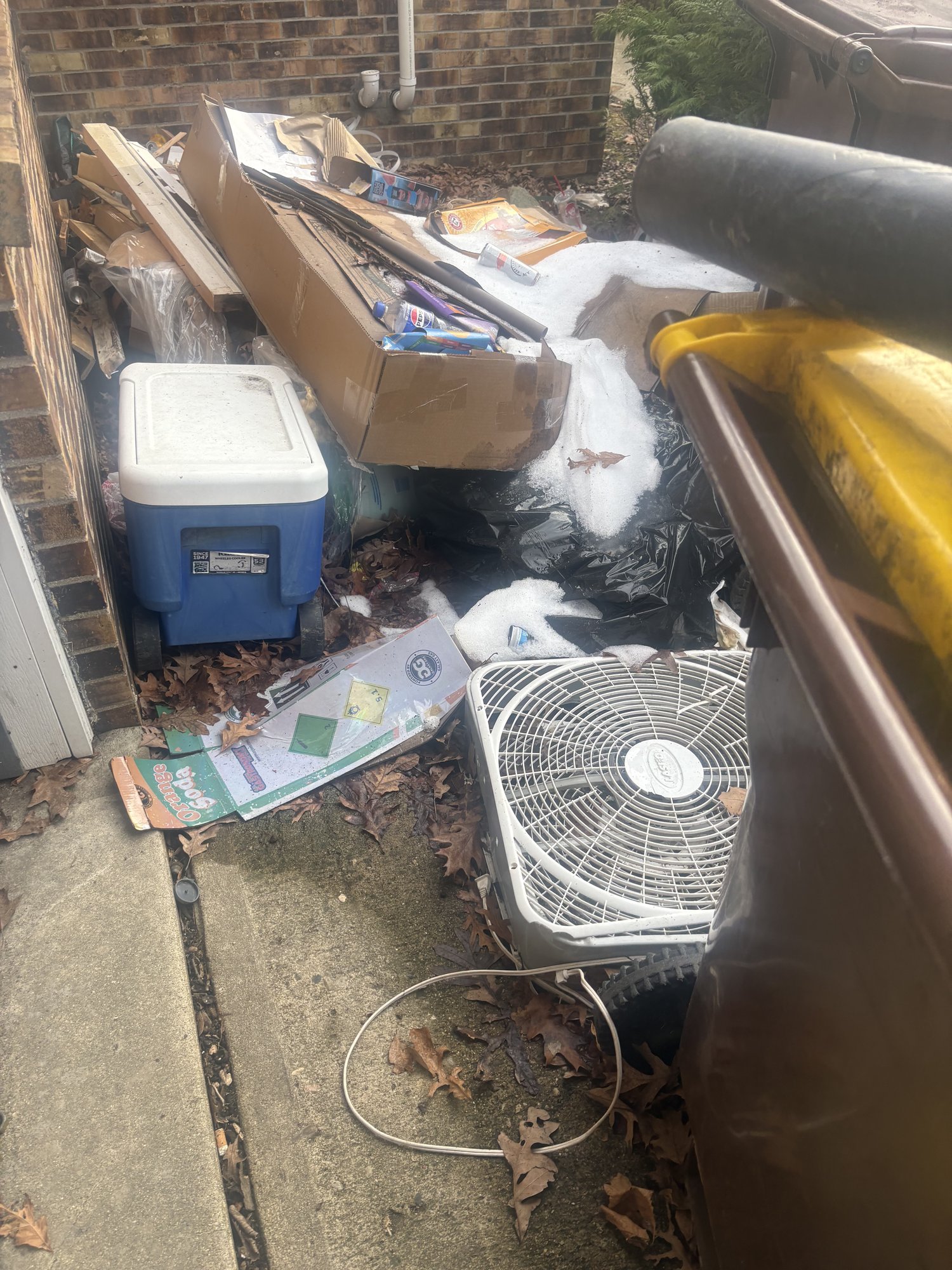 Pile of household junk, cooler, boxes, fan, and bagged debris stacked against a brick wall