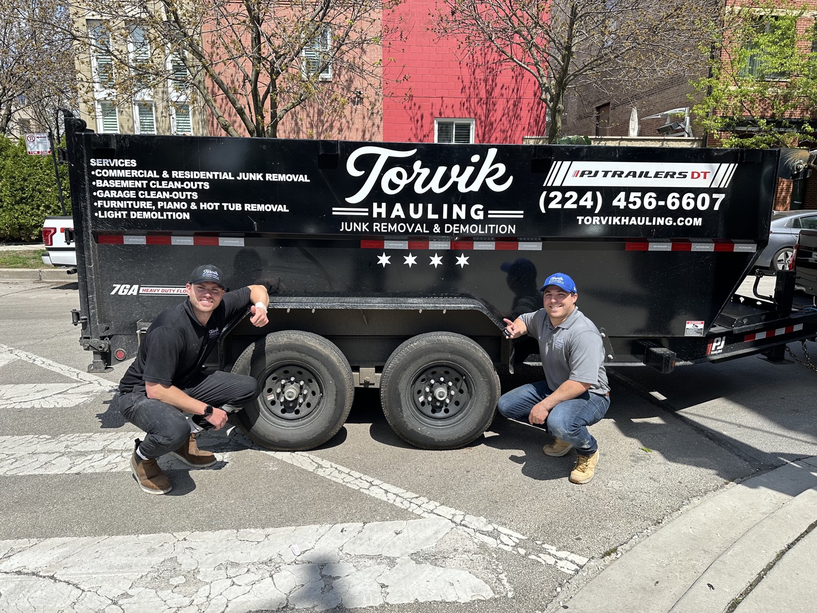 The Torvik Hauling team kneeling beside their branded dump trailer with services list and phone number on the panel