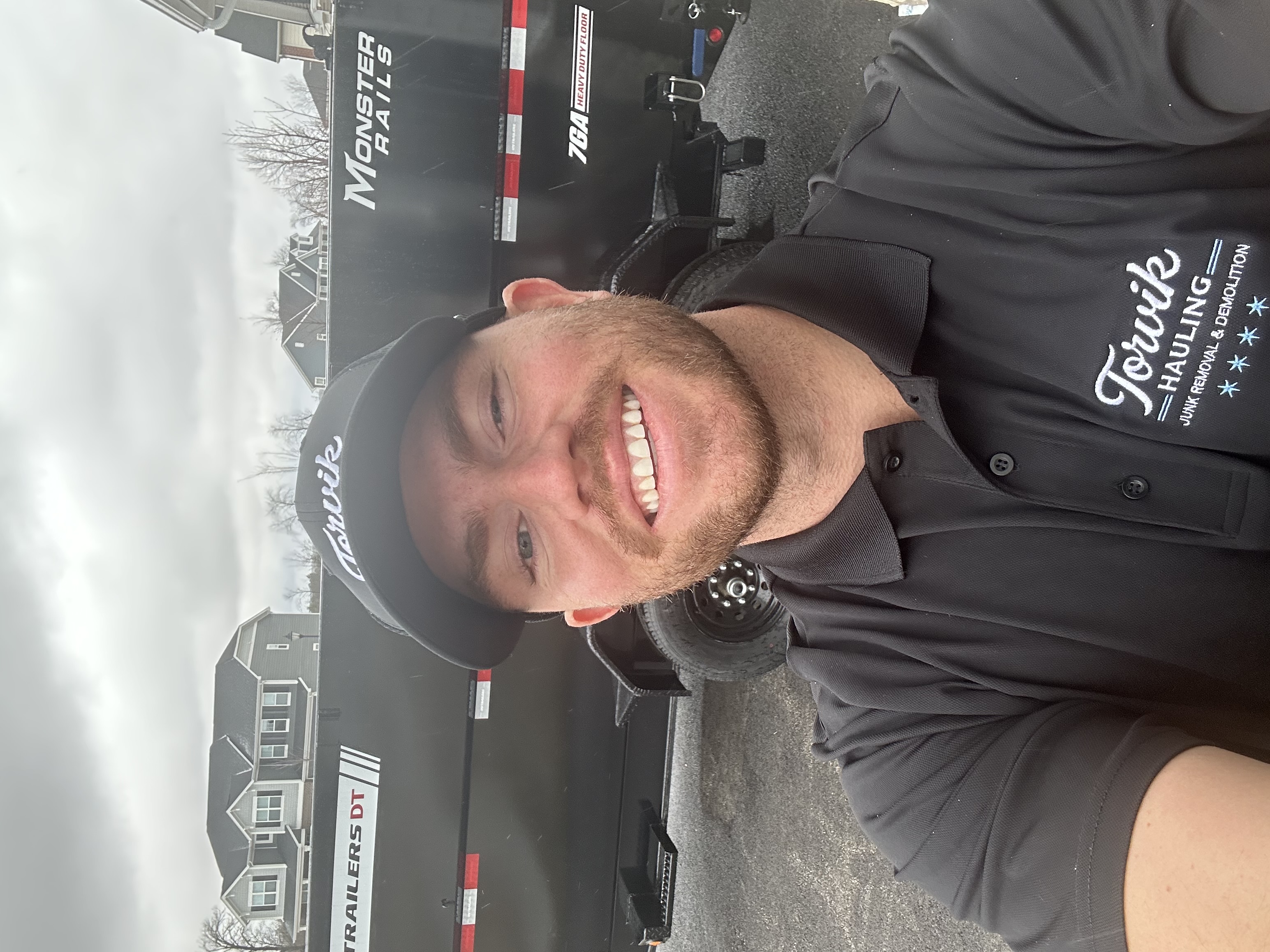 Torvik Hauling owner smiling in a branded black Torvik polo and cap beside the Torvik dump trailer